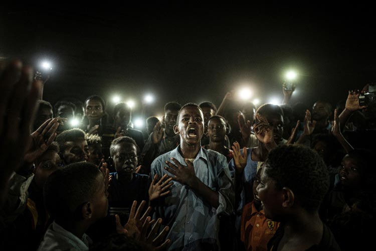 Yasuyoshi Chiba aus Japan wurde für sein Bild vom Aufstand im Sudan mit dem Hauptpreis ausgezeichnet. Ein junger Mann, der von Mobiltelefonen beleuchtet wird, rezitiert Gedichte, während Demonstrant*innen gegen den sudanesischen Diktator protestieren.